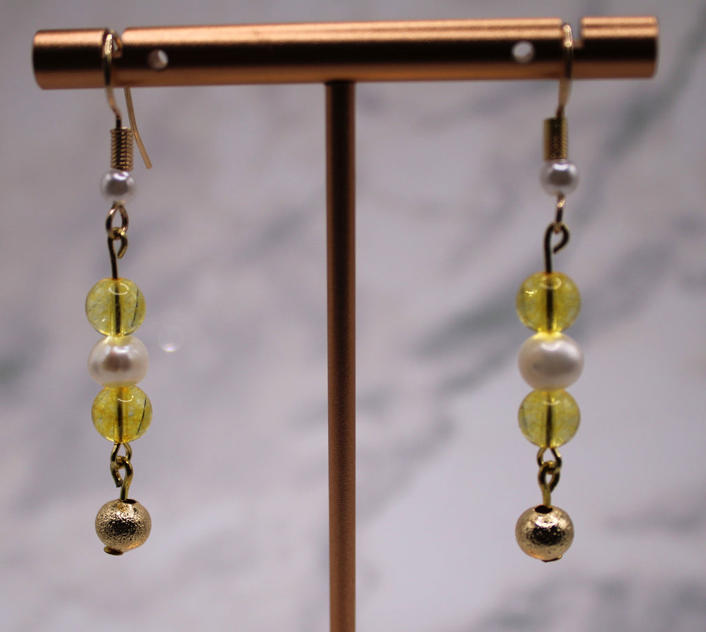 A pair of dangling earrings displayed on a metallic stand. Each earring has a gold fishhook, a small white pearl bead, two translucent yellow beads, and a textured gold ball at the bottom.