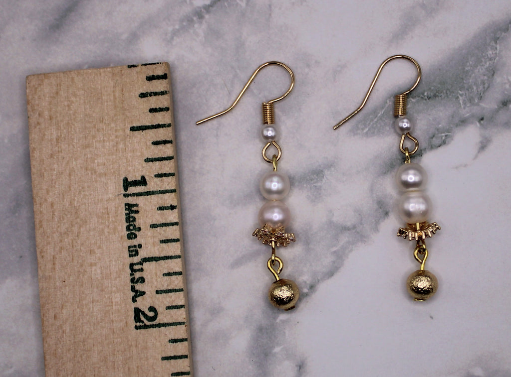 A pair of gold and pearl drop earrings displayed next to a wooden ruler on a white and gray marble surface.