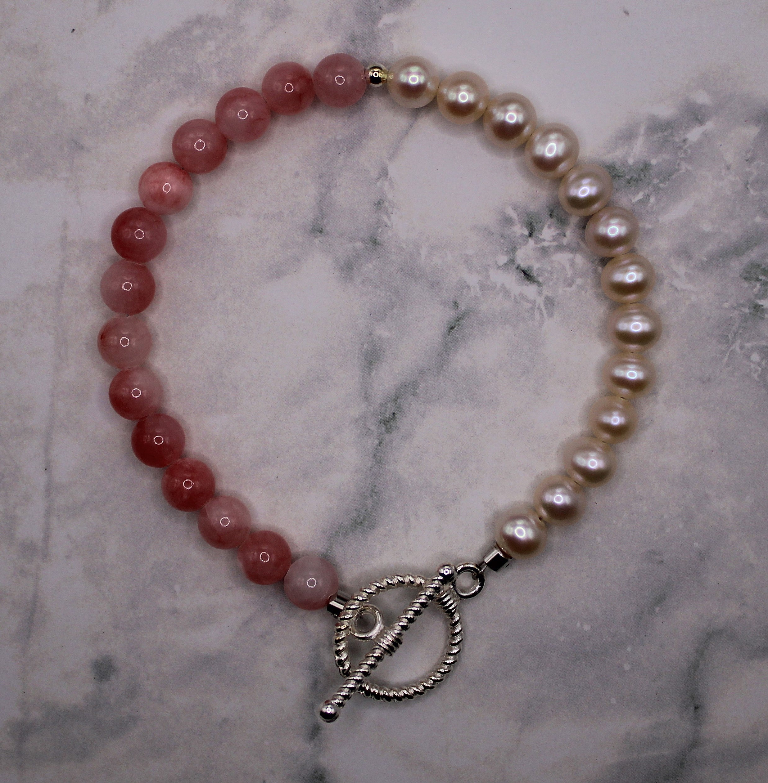A beaded bracelet with alternating pinkish-red and cream pearl-like beads, finished with a silver toggle clasp, displayed on a white and gray marble surface.