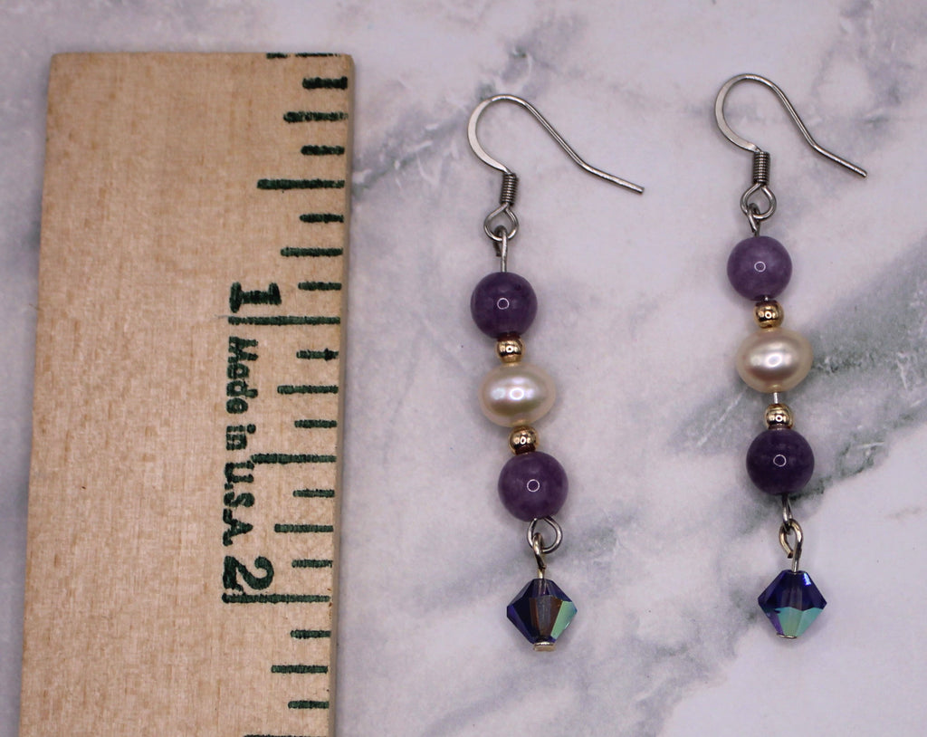 A pair of handmade dangle earrings displayed on a white marble surface next to a wooden ruler for scale. The earrings feature purple beads, a white pearl, a small gold spacer bead, and a faceted dark blue-green iridescent bead at the bottom.